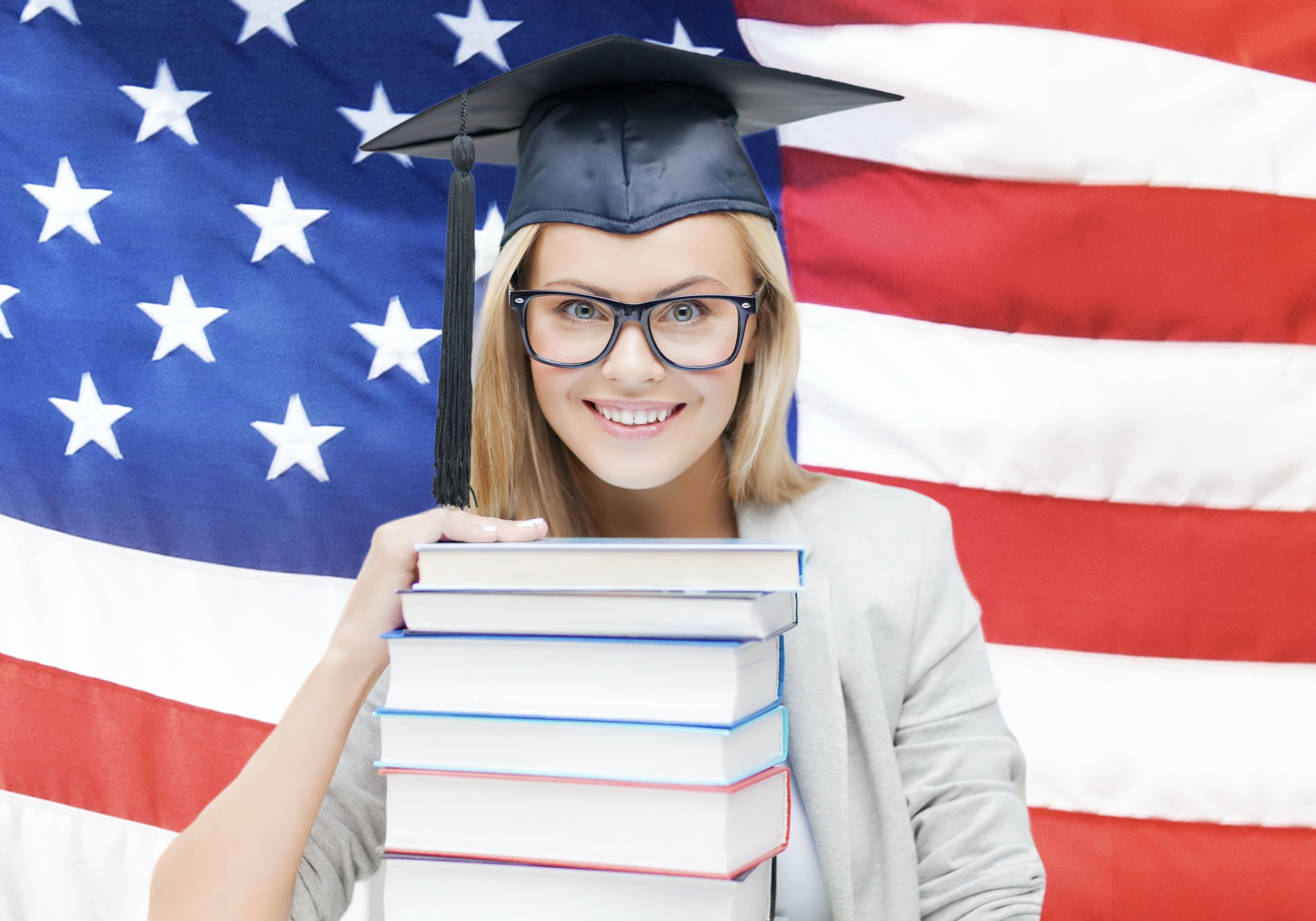 Student in graduation cap holding a stack of books in front of American Flag